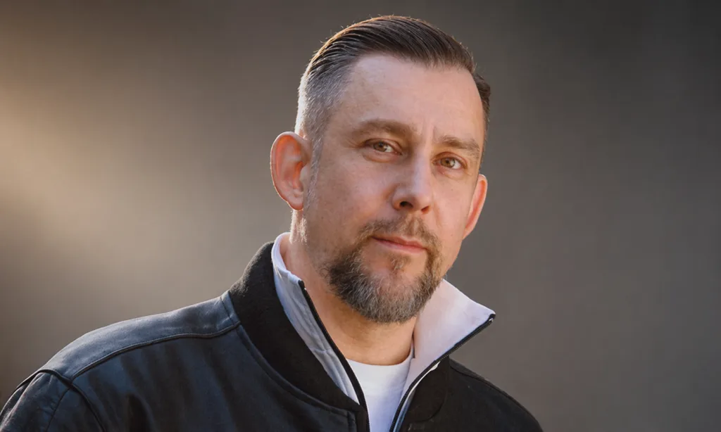 Portrait of a man with short dark hair and a trimmed beard, wearing a black jacket, outdoors with a neutral background.