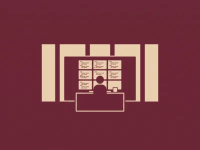 Silhouette of a person sitting at a desk in front of a large monitor with several smaller screens around, suggesting a video conference setup.