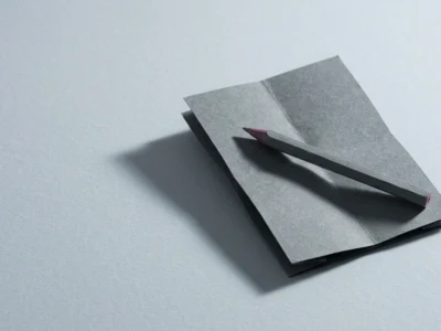 Gray folded paper origami card with a pencil resting across it on a pale blue surface (minimalist stationery scene).