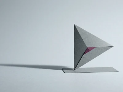 Abstract geometric paper sculpture: gray triangular form on a flat base, with a small pink accent at the center.