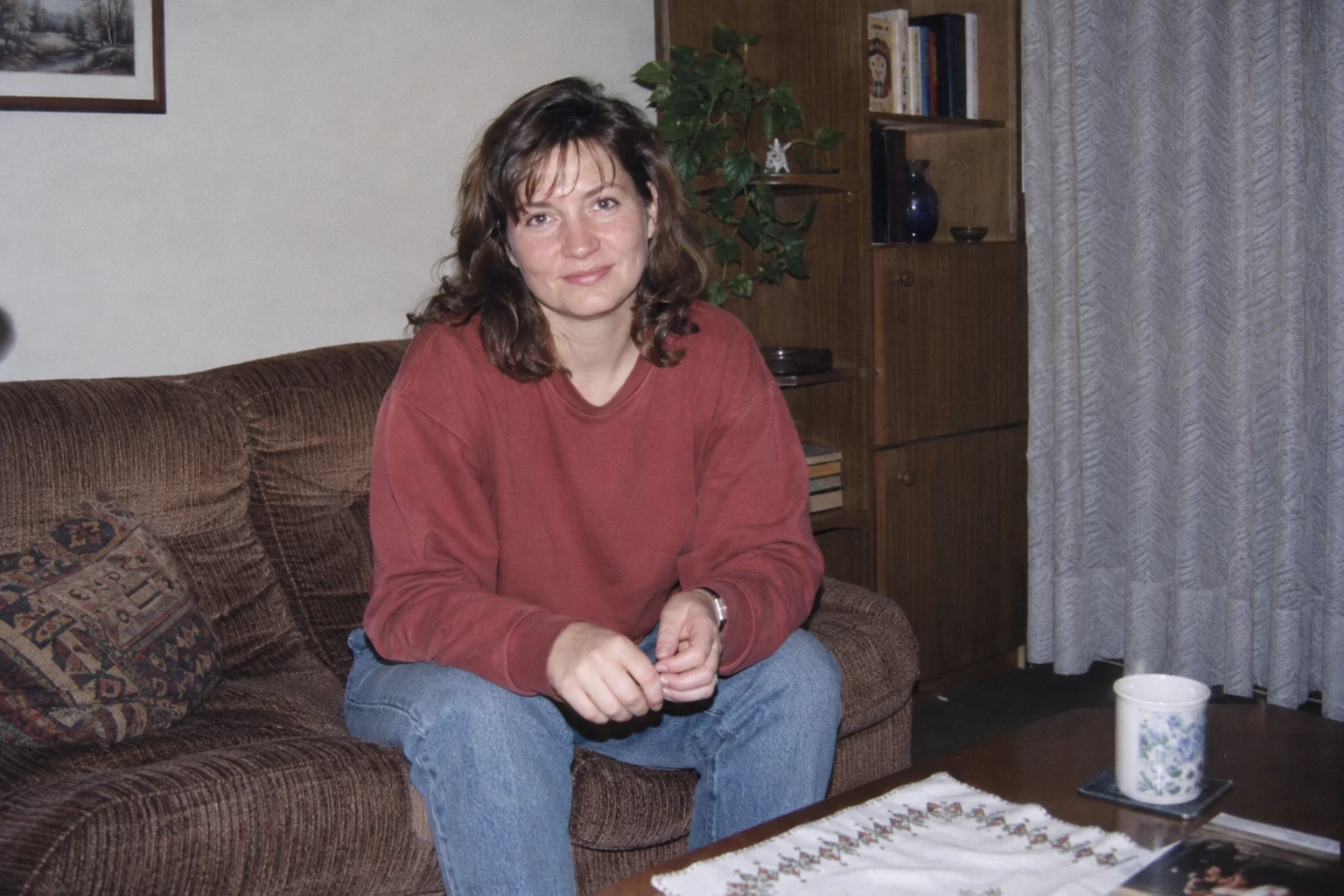 Kobieta w ceglastym swetrze i jeansach na brązowej kanapie w małym pokoju - Promptowy Kobieta w ceglastym swetrze i jeansach na brązowej kanapie w małym pokoju