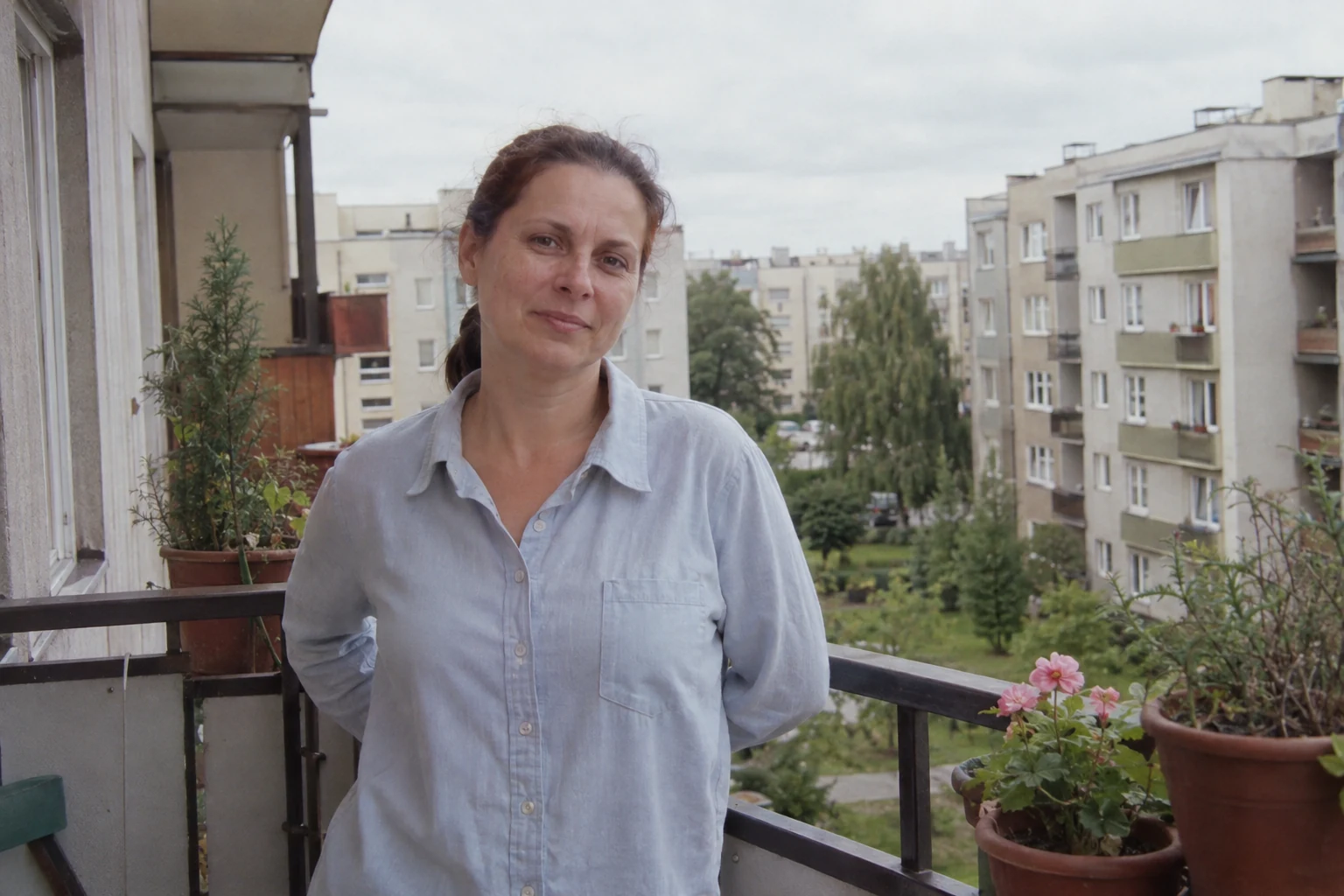 Kobieta w jasnoniebieskiej koszuli na małym balkonie z doniczkami i metalową balustradą - Promptowy Kobieta w jasnoniebieskiej koszuli na małym balkonie z doniczkami i metalową balustradą