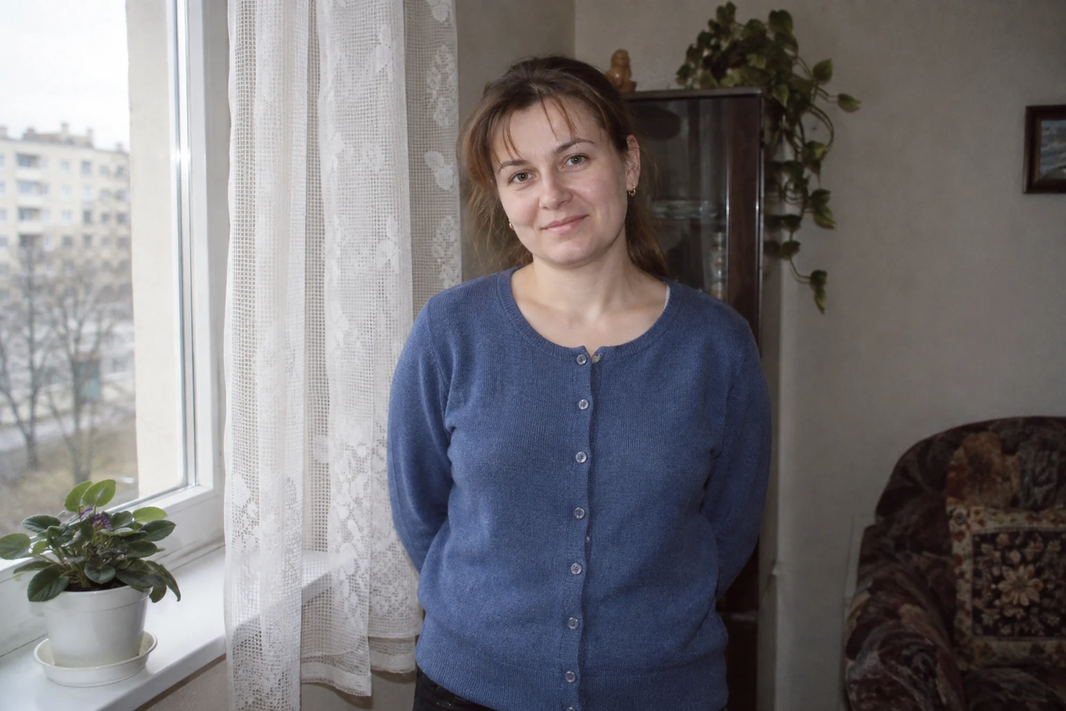 Kobieta w niebieskim kardiganie przy oknie z firanką i doniczką na parapecie - Promptowy Kobieta w niebieskim kardiganie przy oknie z firanką i doniczką na parapecie