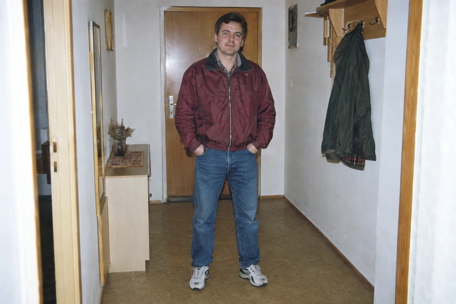 Mężczyzna w bordowej kurtce w przedpokoju z drewnianymi drzwiami i wieszakiem - Promptowy Mężczyzna w bordowej kurtce w przedpokoju z drewnianymi drzwiami i wieszakiem