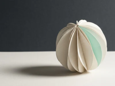 White and pale green paper origami ball folded into concentric petals, resting on a light surface with a soft shadow behind it.