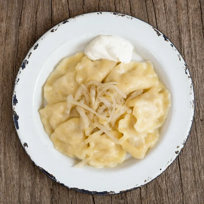 Pierogi na wyszczerbionym emaliowanym talerzu, z cebulką i kleksem śmietany, drewniany blat, miękkie światło dnia.
