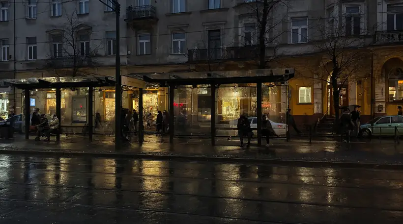 Deszczowy przystanek tramwajowy o zmierzchu: odbicia na mokrym asfalcie, ciepłe witryny, dokumentalny charakter.
